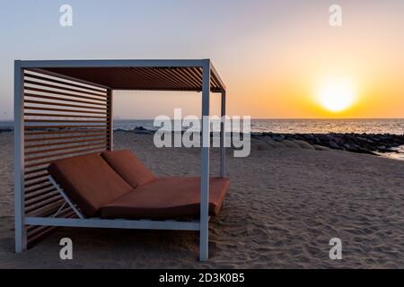 Leere Doppel-Sonnenliege am Sandstrand in Dubai, mit orangefarbenem Sonnenuntergang im Hintergrund und ruhigem Meer. Stockfoto