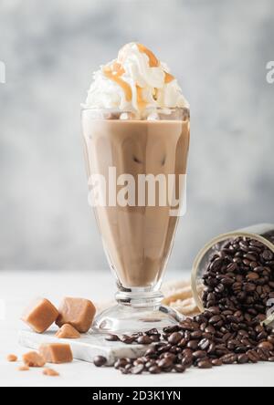 Schwarzer Eiskaffee mit frischer Milch und Karamell mit Sahne in klassischem Milchshake-Glas und Glas roher Kaffeebohnen auf hellem Tischhintergrund. Stockfoto