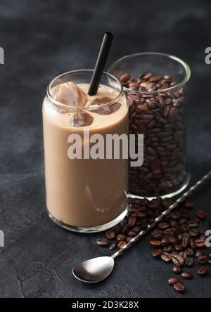 Glas eisgekühlter Kaffee und Milch mit freien rohen Kaffeebohnen in Glasbehälter mit langem Löffel auf schwarzem Hintergrund. Stockfoto
