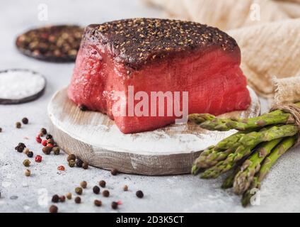 Scheibe Roher Rinderoberseite mit Salz und Pfeffer auf runder Hackbrett mit Spargelspitzen und Knoblauch auf hellem Küchentisch. Stockfoto