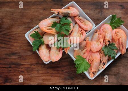 Gekochte rosa Garnelen in der Schale mit Petersilie serviert. Muschelgarnelen auf den weißen Tellern auf dem rustikalen Holztisch. Draufsicht, Kopierbereich Stockfoto