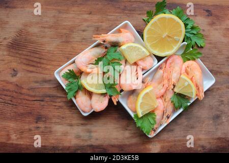 Gekochte rosa Garnelen in der Schale mit Petersilie und Zitrone serviert. Muschelgarnelen auf den weißen Tellern auf dem rustikalen Holztisch. Draufsicht Stockfoto