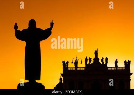 Hintergrundbeleuchtung der Fassade der Basilika San Giovanni, mit der Statue des Heiligen Franziskus von Assisi im Vordergrund, bei Sonnenuntergang in Rom, Italien. Stockfoto