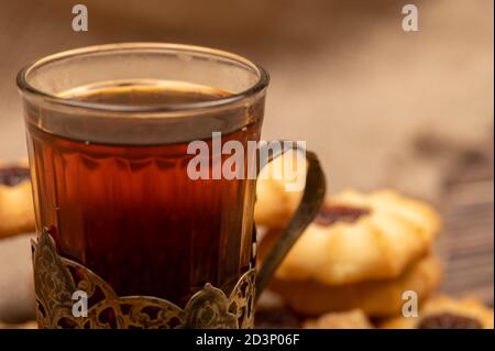 Hausgemachte Gebäck-Kekse mit Marmelade und einem facettierten Glas Tee in einem Vintage-Cup-Halter auf einem Hintergrund von homespun Stoff mit einer rauen Textur, close-up, Stockfoto