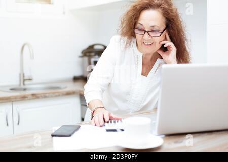 Pensionierte Frau, die sich auf dem Küchentisch ausruhte. Sie verwendet einen Rechner und prüft Rechnungen. Stockfoto