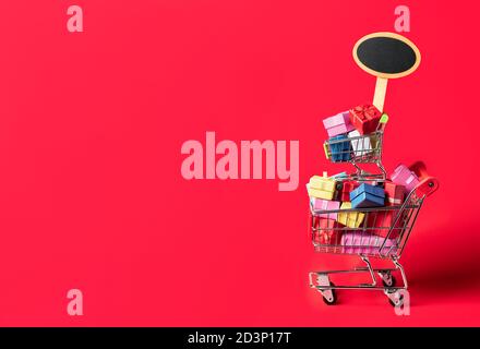 Warenkorb voller farbenfroher Geschenke und leerem Plakat, isoliert auf rotem Hintergrund. Black Friday Verkaufskonzept. Einkaufen von Weihnachtsgeschenken. Stockfoto