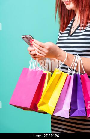 Frau, die Einkaufstaschen an den Händen hält und ein Smartphone benutzt. Nahaufnahme einer rothaarigen Frau, die mit bunten Papiertüten einkaufen. Stockfoto