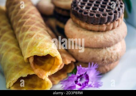 Waffelbrötchen und Kekse auf einem weißen Teller und eine Tasse Tee. Waffeln auf der Seite. Komposition auf einem hölzernen Hintergrund. Sichtbare Waffelstruktur Stockfoto