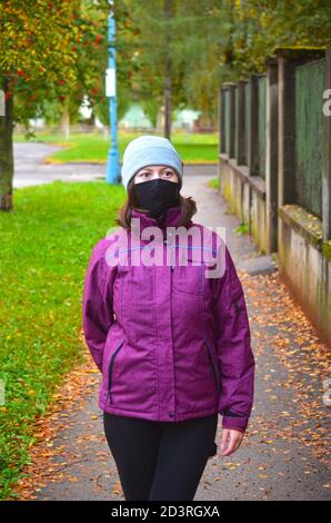 Junge Frau in warmen Kleidern tragen lila Jacke mit schwarzen wiederverwendbaren Gesichtsmaske und stricken Hut stehen auf Bürgersteig mit braunen Blättern bedeckt. Herbst Stockfoto