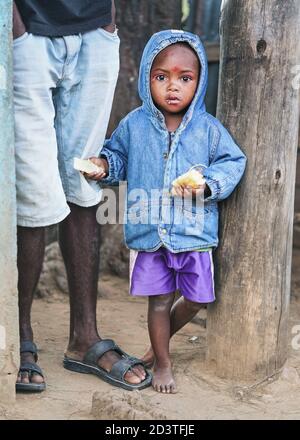 Ranohira, Madagaskar - 29. April 2019: Unbekannter madagassischer Junge, in blauer Jeansjacke mit Kapuze und Shorts, barfuß, neben den Beinen seines Vaters stehend, ho Stockfoto