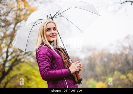 Schöne blonde Frau in hellen Purpurmantel und Schal halten transparenten Regenschirm im Herbst Park Hintergrund, regnerisches Wetter, Spaziergang im Herbst Saison Stockfoto