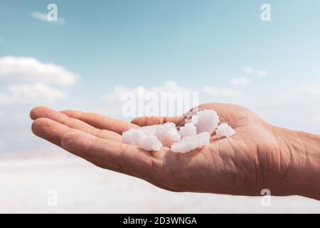 Hand hält Bündel von rosa weißen Salzflocken Kristalle über Wasser mit blau verschwommenem Himmel Hintergrund. Spa Resort sonnige Nahaufnahme Stockfoto