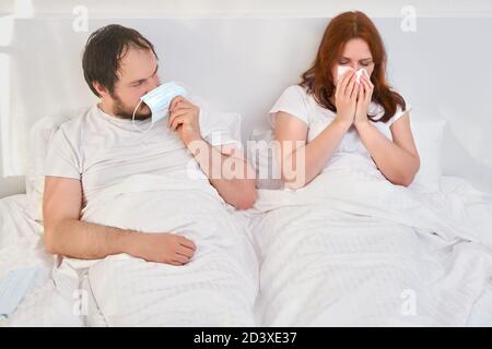 Ein Mann wird durch eine medizinische Maske vor einer kranken Frau geschützt, die zusammen in einem weißen Bett liegt, Konzept. Beziehungsprobleme gepaart mit Isolation durch Cor Stockfoto