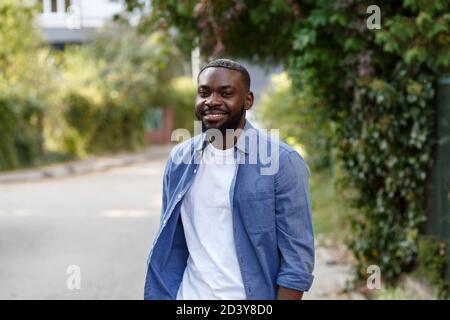 Nahaufnahme eines glücklichen afroamerikanischen Mannes. Schöner junger Mann in blauem Denim-Hemd, der auf der Straße spazierengeht und lächelt Stockfoto
