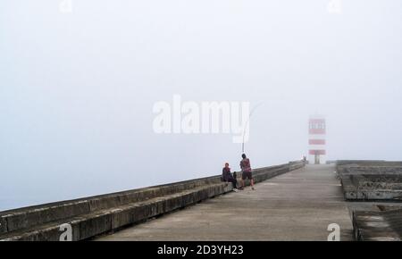 Frau und Mann fischen entlang der Wellenbrecher von Farolins da Barra do Douro, in einem nebligen Morgen. Porto, Portugal Stockfoto