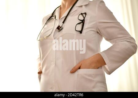 Der Arzt steht, beide Hände in der Tasche, ein Stethoskop hängt um ihren Hals, eine Brille mit dunklem Rahmen auf ihrer Brust. Stockfoto