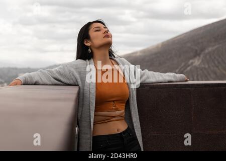 Die junge Lateinerin lehnt sich mit geschlossenen Augen und dem Kopf zurück an einen Handlauf, trägt ein orangefarbenes Oberteil und Jeans, im Hintergrund Berge Stockfoto