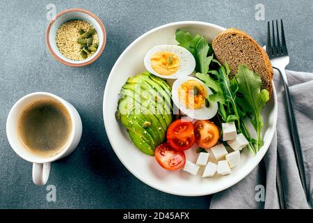 Gesunde vegane Frühstückschüssel. Avocado, Ei, Tomate, Feta-Käse, Rucola und Brot und eine Tasse Kaffee. Draufsicht Stockfoto