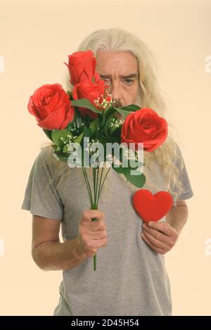 Studio shot der älteren bärtigen Mann versteckt sich hinter roten Rosen halten roten Herzen zum Valentinstag bereit Stockfoto