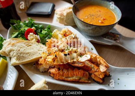 Kaisergranat wird auf einem weißen, fischförmigen Teller gegrillt. Stellen Sie wunderschöne Brote und Gemüse auf den Esstisch. Es gibt eine Schüssel Suppe auf der Seite. A Stockfoto