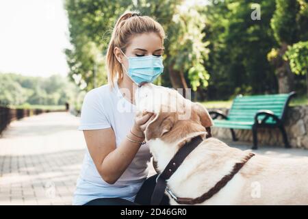 Schöne blonde Mädchen und ihr labrador sind umarmen in ein parken, während Sie eine medizinische Maske tragen Stockfoto