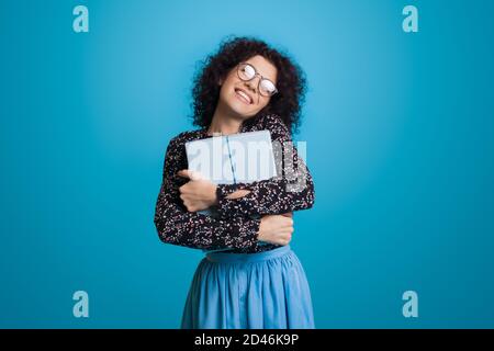 Süße kaukasische Frau mit lockigen Haaren und Brille umarmt Eine Geschenk-Box lächelt auf einer blauen Studiowand in Ein Kleid Stockfoto