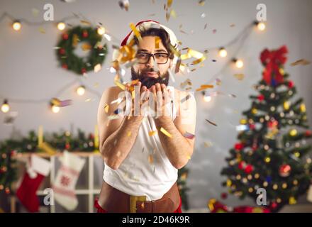 Fröhlicher junger Mann in Santa Hut weht Glitter Konfetti, wünschen Liebe und Glück im neuen Jahr Stockfoto