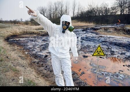 Wissenschaftler tragen weiße Schutzhülle und Gasmaske neben einem Biogefahrenschild an kontaminiertem Wasser stehen. Betretet keine gefährliche Zone, haltet Euch fern, der Mann zeigt zur Seite Stockfoto