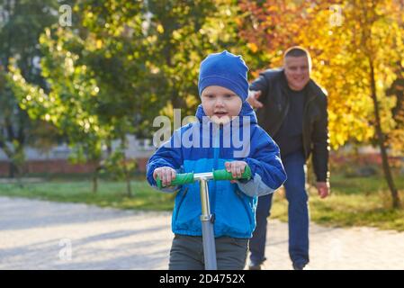 Papa lehrt seinen zweijährigen Sohn, im Herbst einen Roller im Park zu fahren. Stockfoto