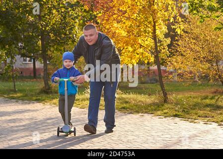 Papa lehrt seinen zweijährigen Sohn, im Herbst einen Roller im Park zu fahren. Stockfoto