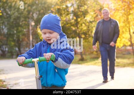 Papa lehrt seinen zweijährigen Sohn, im Herbst einen Roller im Park zu fahren. Stockfoto