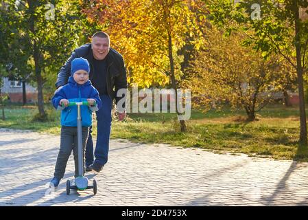 Papa lehrt seinen zweijährigen Sohn, im Herbst einen Roller im Park zu fahren. Stockfoto