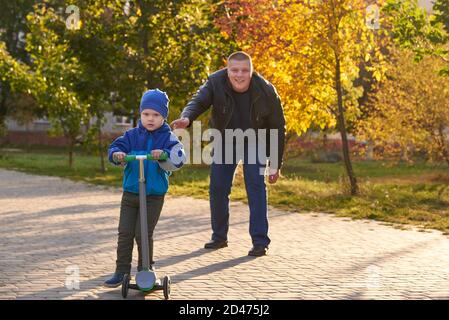 Papa lehrt seinen zweijährigen Sohn, im Herbst einen Roller im Park zu fahren. Stockfoto