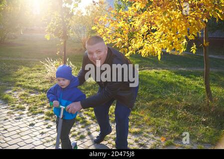 Papa lehrt seinen zweijährigen Sohn, im Herbst einen Roller im Park zu fahren. Stockfoto