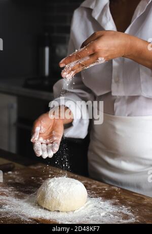 Blick auf den Schreibtisch des Kochs. Das Foto zeigt die Hände des Kochs, der Mehl auf einen Teig streut. Stockfoto