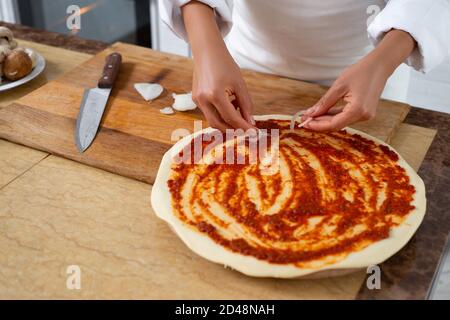 Blick auf die Hände eines Küchenchefs, der beginnt, Zwiebeln auf einem Stück Kochpizza zu legen. Stockfoto