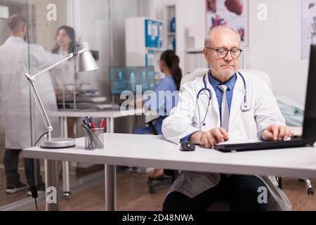 Arzt mit grauen Haaren in Klinik Schrank arbeiten am Computer und junge Sanitäter diskutieren mit Frau Patient auf Krankenhausflur. Stockfoto