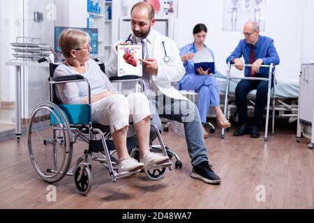 Medizinisches Personal, das eine Broschüre mit Arrhythmien an behinderte ältere Frau im Rollstuhl präsentiert. Mann mit Behinderungen, Gehrahmen sitzt im Krankenhausbett. Gesundheitssystem, Klinikpatienten. Stockfoto