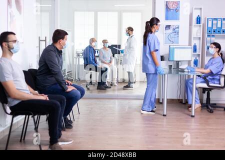 Krankenschwestern tragen Gesichtsmaske gegen Coronavirus und ein Gespräch auf Krankenhaus Wartebereich. Assistent, der am Empfangscomputer arbeitet. Doktor, der sich mit einem älteren Paar ausschälte Stockfoto