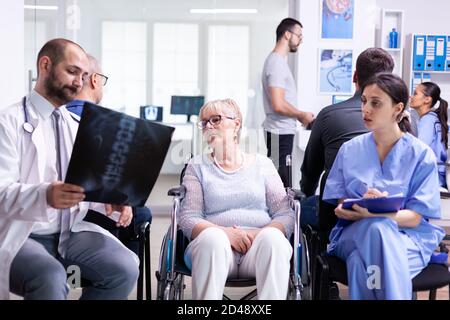 Arzt mit Stethoskop hält behinderte ältere Frau Radiographie im Rollstuhl, während mit ihr im Krankenhaus Wartebereich sprechen. Patient fragt nach seinem Termin an der Klinikrezeption. Stockfoto