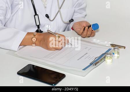 Indische Dame Arzt schreibt eine ärztliche Verschreibung Form mit einem Medikamentenakte mit Impfstoffflaschen auf dem Tisch Stockfoto