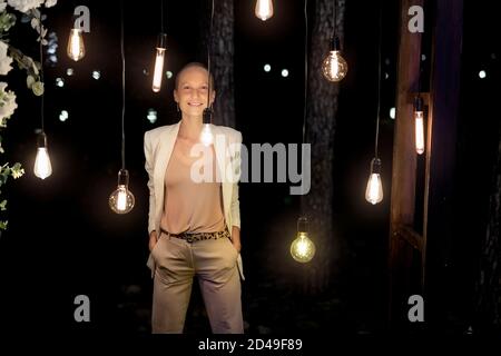 Porträt der jungen erwachsenen kaukasischen Glatze Frau lächeln und stehen unter vielen edison Glühbirnen am Wald im Freien gehängt. Die richtige Lösung Stockfoto