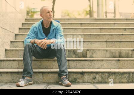 Voller Körper geschossen des Kahlen älterer Mann denken, während auf der Treppe sitzen in Bangkok, Thailand Stockfoto