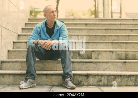 Voller Körper geschossen des Kahlen älterer Mann denken, während auf der Treppe sitzen in Bangkok, Thailand Stockfoto