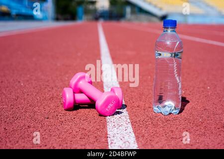 Sportgerät und -Ausrüstung. Kurzhanteln auf der Rennstrecke im Stadion. Hydratation nach der Fitness. Gesunder Lebensstil. Wasserhaushalt im Körper beim Training. Langhanteln und Flasche auf der Laufstrecke. Stockfoto