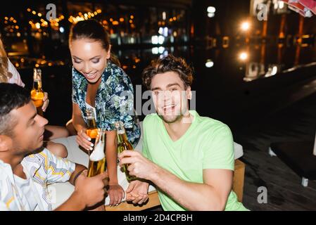 Selektiver Fokus des jungen Mannes beim Anklirren auf die Kamera Bier mit Freunden in der Nacht Stockfoto