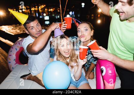 Selektiver Fokus von jungen Freunden in Party Caps Spaß haben Und halten Einweg-Tassen in der Nacht Stockfoto