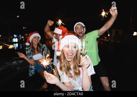 Selektive Fokus der Frau in santa Hut hält Glas Champagner und Wunderkerzen in der Nähe von Freunden in der Nacht Stockfoto