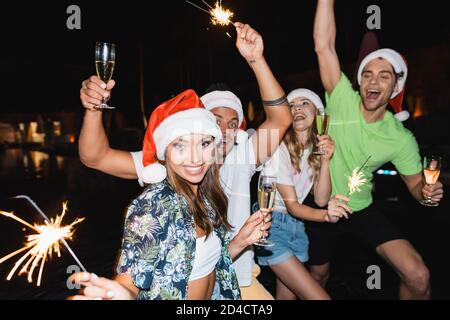 Selektive Fokus der jungen Frau in santa Hut hält Glas Champagner und Wunderkerzen in der Nacht Stockfoto