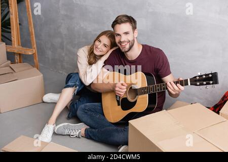 Zufrieden Frau umarmt Freund spielen akustische Gitarre in der Nähe von Boxen, Relocation-Konzept Stockfoto